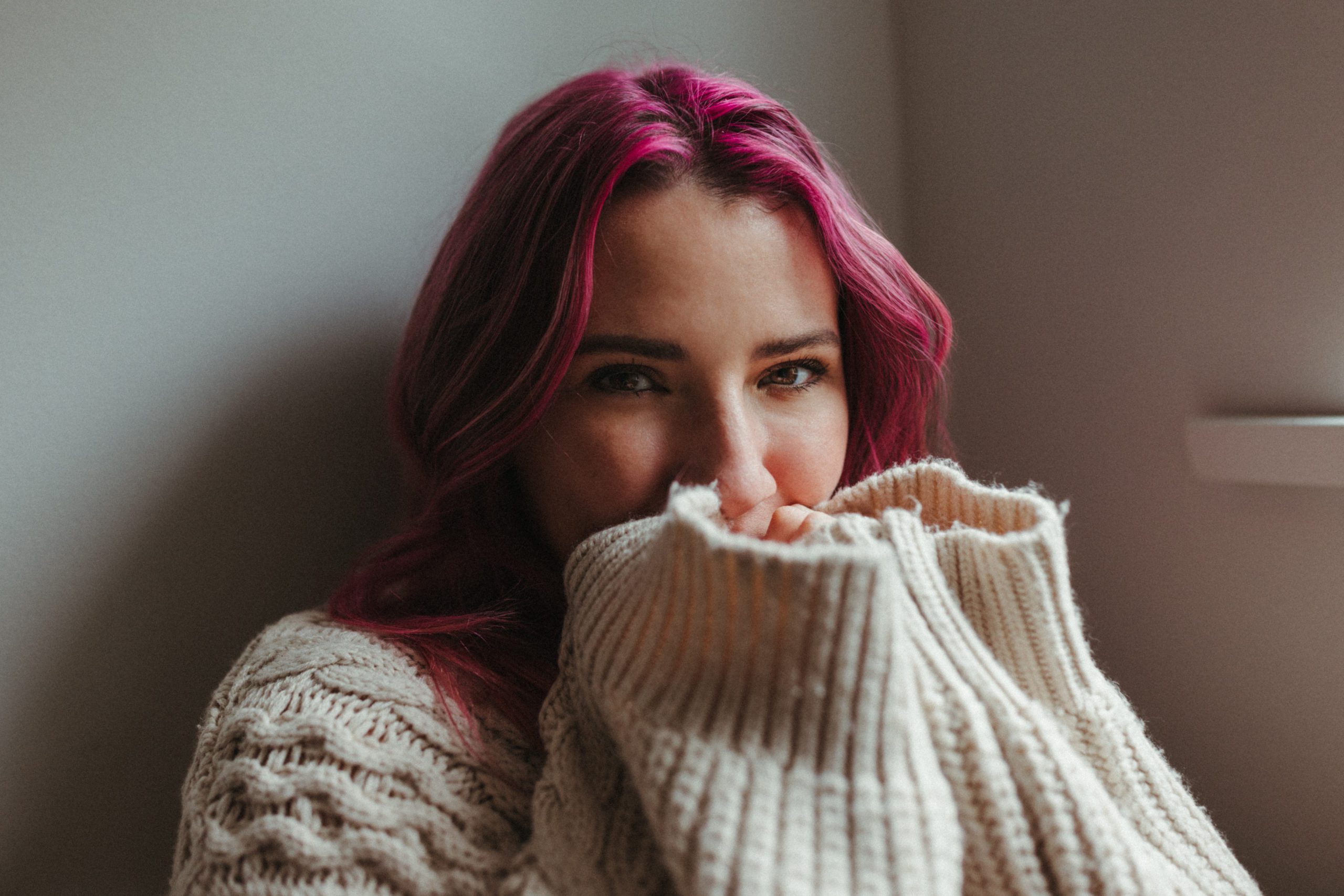 Young girl with pink hair holding her hands up to her face in a long sleeve sweater